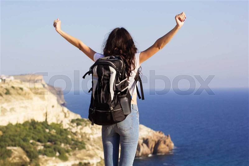 Back view of young pretty slim woman standing with backpack on cliff\'s edge and looking into sea, stock photo