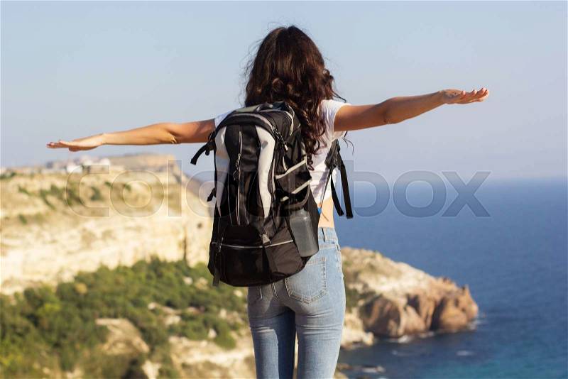 Back view of young pretty slim woman standing with backpack on cliff\'s edge and looking into a wide valley, stock photo