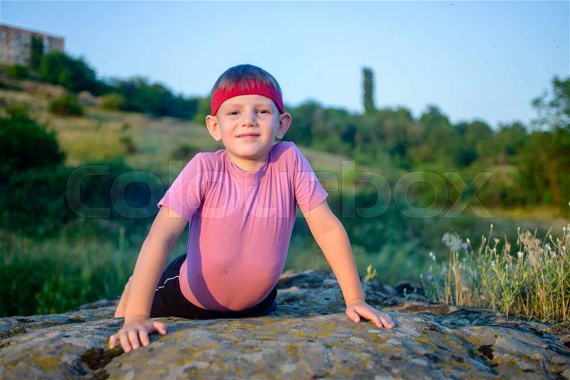 Cute little boy trying to do press-ups as he works out on a rock looking up at the camera with a wry smile, fitness and active lifestyle concept, stock photo