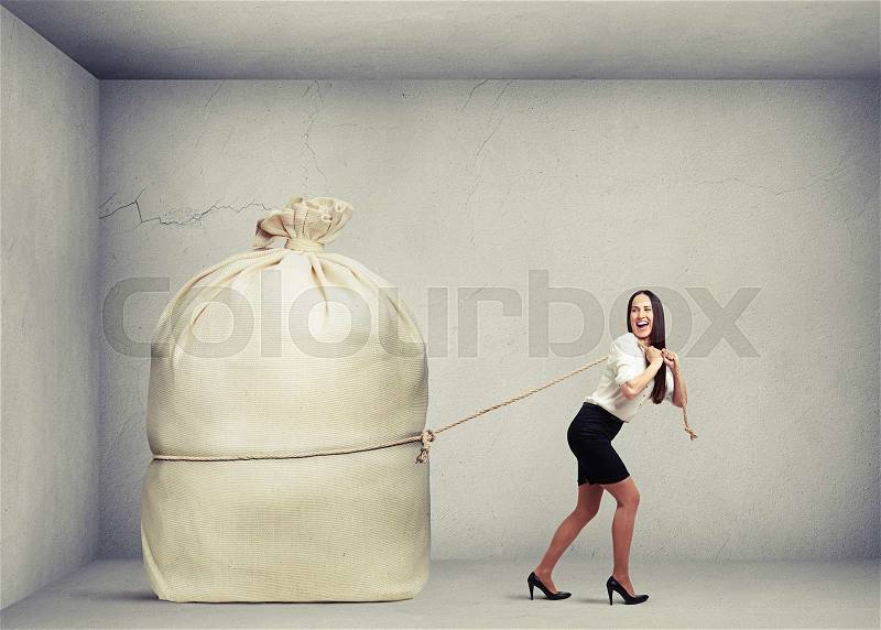 Happy woman in formal wear pulling big bag in grey concrete room, stock photo