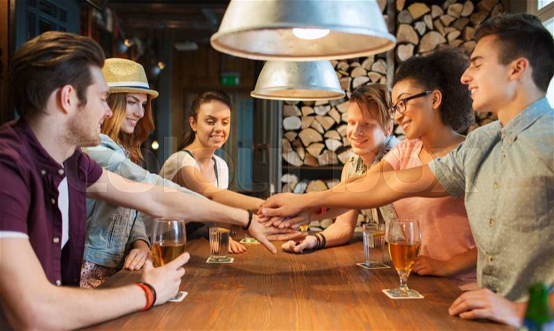 People, leisure, friendship and gesture concept - group of happy smiling friends with drinks putting hands on top of each other at bar or pub, stock photo
