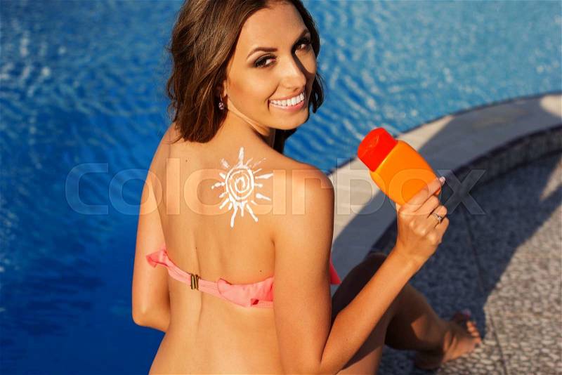 Sexy smiling girl with sun shape drawing from sunscreen lotion on her shoulder is resting near swimming pool with blue water, tropical resort, stock photo