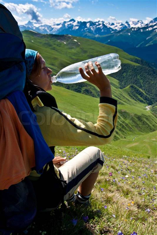 Hiker girl drinking water after hard rush and relaxing on the meadow, with mountains background, stock photo