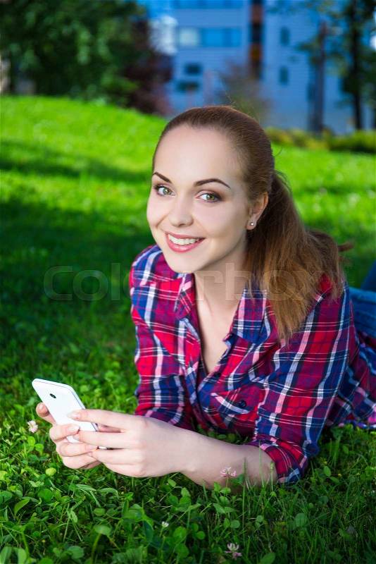 Smiling teenage girl lying with smart phone on grass in summer park, stock photo