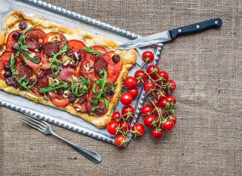 Homemade square pizza with meat, salami, cherry-tomatoes and fresh arugula on a silver tray over a sackcloth background, stock photo