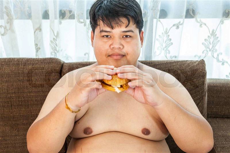 Asian fat man eating hamburger seated on armchair, stock photo