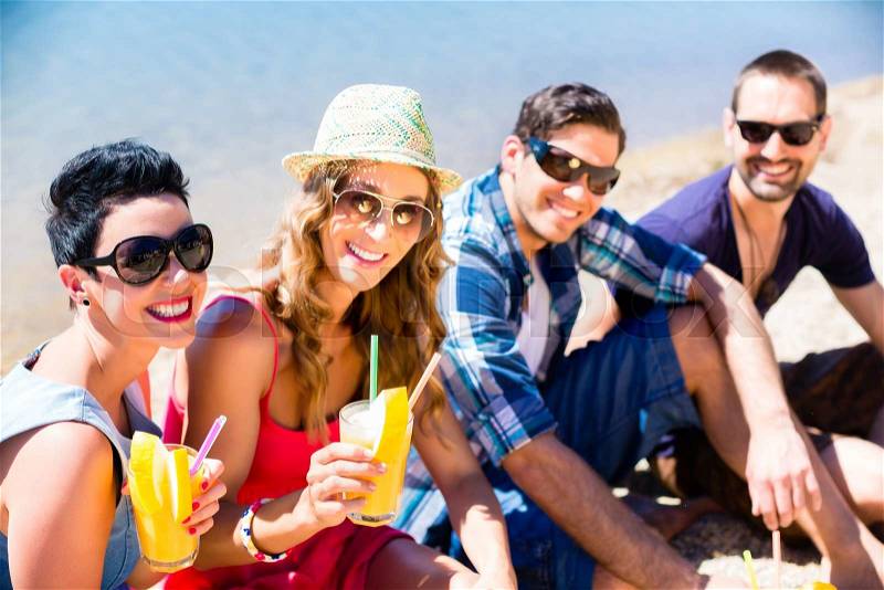 Four friends sitting on lake beach with cocktails in the summer sun , stock photo