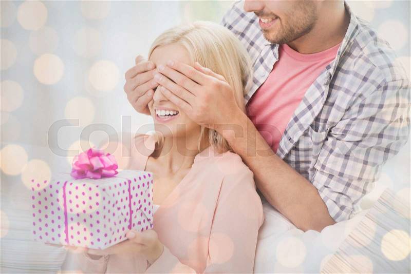 Relationships, love, people, birthday and holidays concept - happy man covering woman eyes and giving gift box over lights background, stock photo
