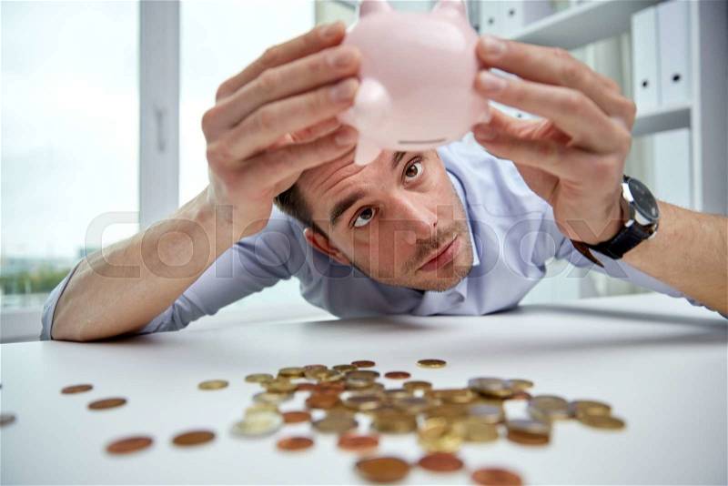 Business, people, finances, crisis and money saving concept - businessman shaking piggy bank and getting coins at office, stock photo