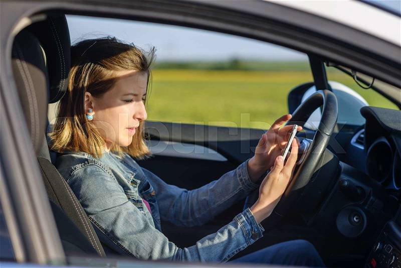 Dangerous female driver reading a text message on her smartphone and taking her attention off the road, profile view, stock photo