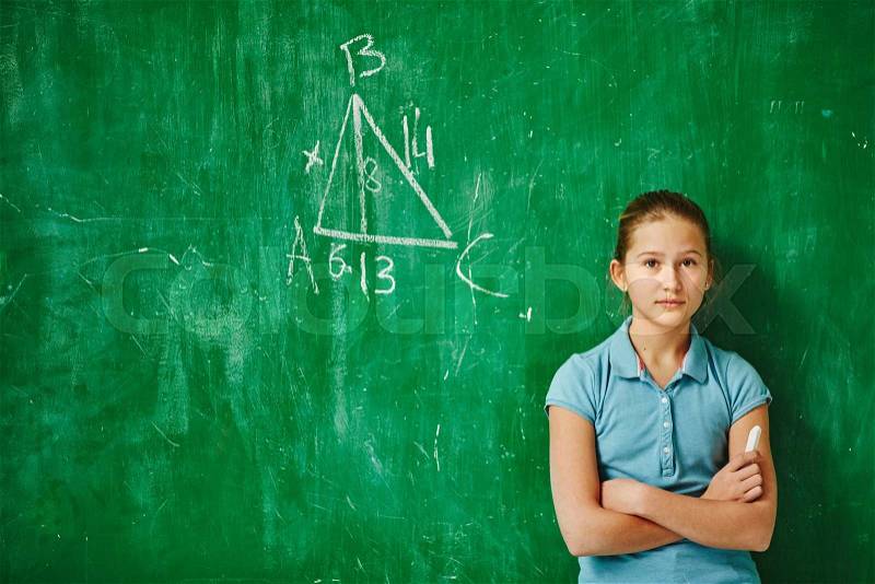 Cute schoolgirl looking at camera while standing by blackboard with geometric task on it, stock photo