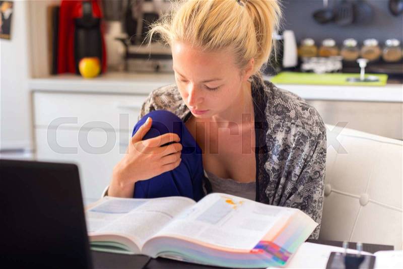 Adult woman in her casual home clothing working and studying remotly from her small flat in the morning. Home kitchen in the background. Great flexibility of web-based courses and study programmes, stock photo