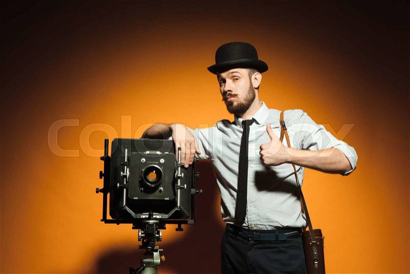 Young positive man in hat as photographer with retro camera on an orange background, stock photo