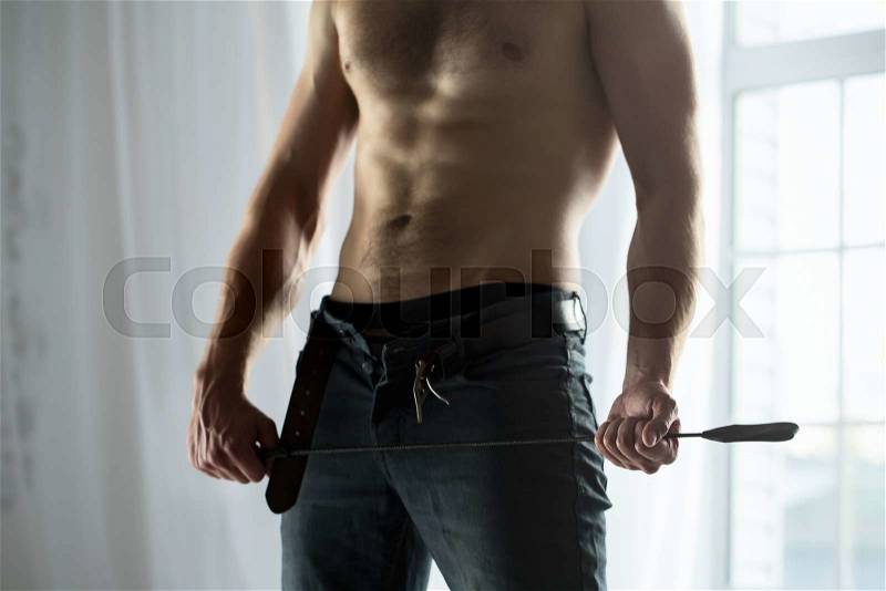 Cropped hands of topless guy with jeans unbuttoned playfully holding a whip BDSM. The picture in the studio on a background window, stock photo