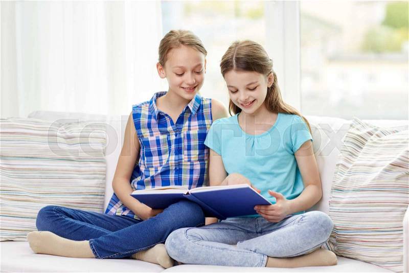 People, children, friends, literature and friendship concept - two happy girls sitting on sofa and reading book at home, stock photo