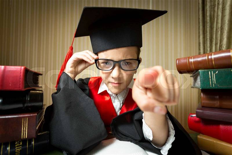 Funny portrait of girl in eyeglasses and graduation cap pointing at camera with finger, stock photo