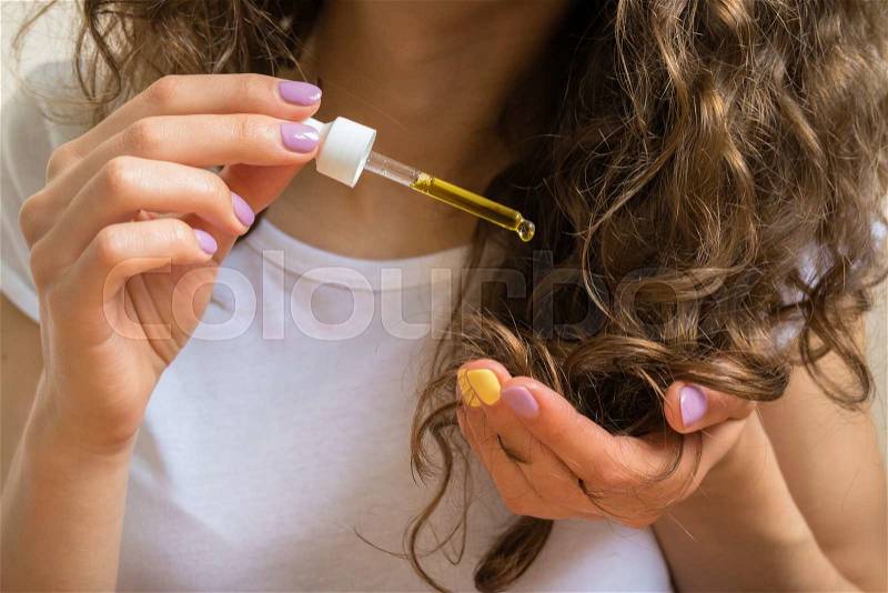 Young girl in white t-shirt holds in her left hand the tips of her curly hair and her right hand applies them yellow oil, stock photo