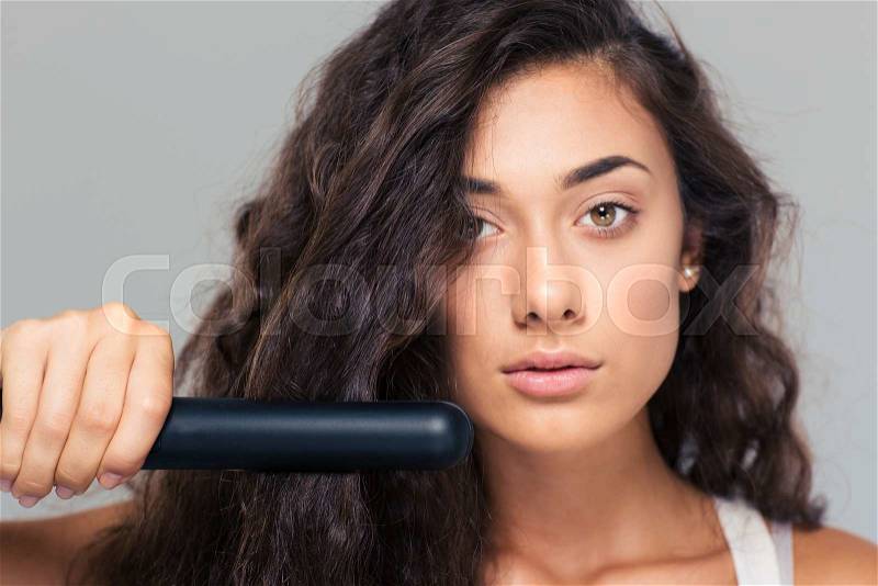 Closeup protrait of a cute woman doing hairstyle with hair straightener over gray background. Looking at camera, stock photo