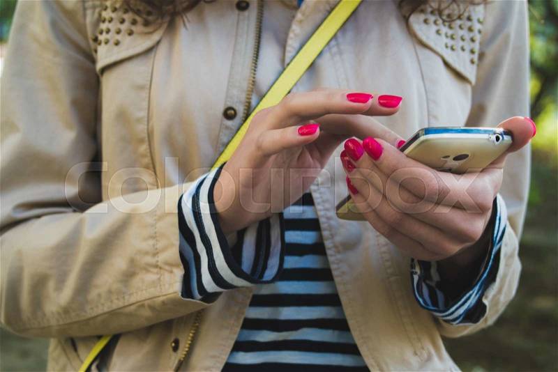 The girl uses the phone close up, she is dressed in a beige jacket and striped shirt, at the shoulder yellow strap from the bag. She holds a white phone with two hands with red nail Polish, stock photo