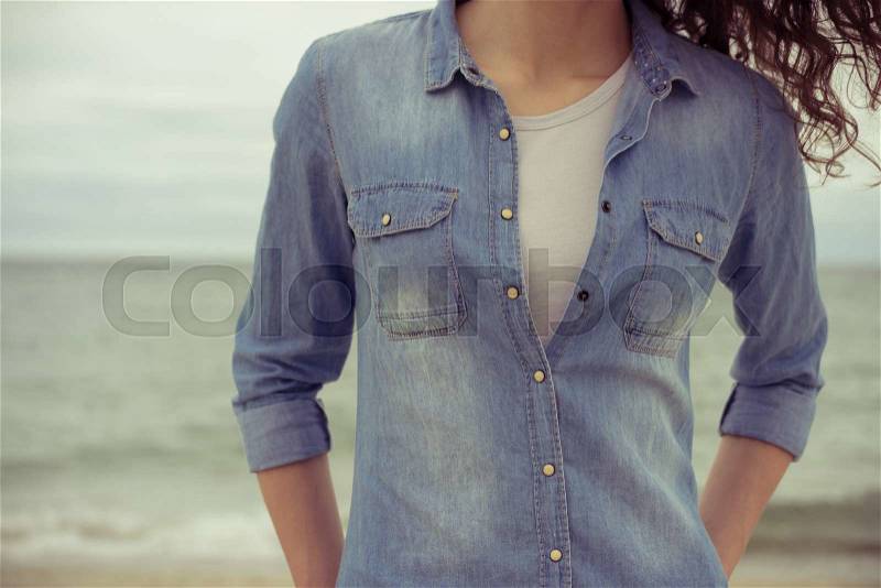Slim woman in a denim shirt and white t-shirt stands on the beach against the sea in cloudy weather. She has curly hair. Her hands in her pockets. Retro colors. Closeup. Relaxed mood, stock photo