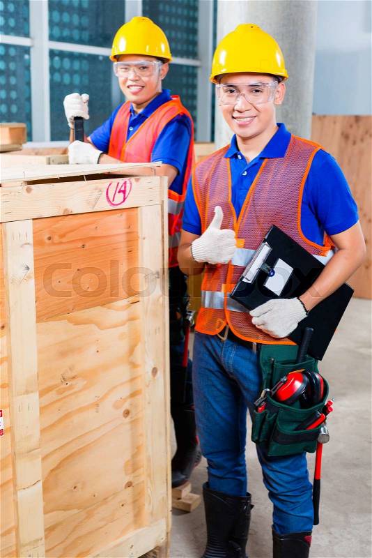 Two Asian Indonesian industrial or construction workers controlling with a checklist a delivery on a tower building site and open a wood box or cargo container with a crowbar , stock photo