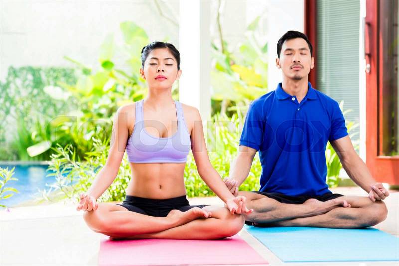 Asian yoga couple in lotus seat mediating in their tropical home in front of garden, stock photo