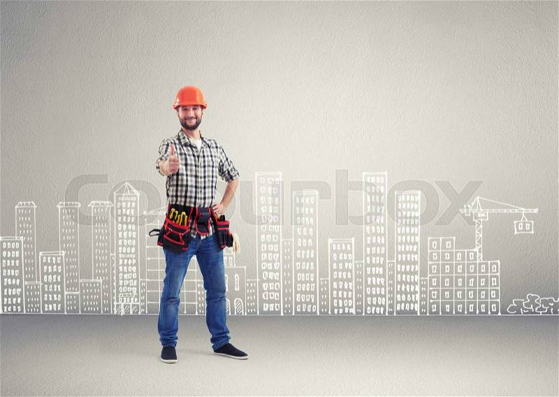 Cheerful builder showing thumbs up and looking at camera over grey wall with drawing cityscape, stock photo