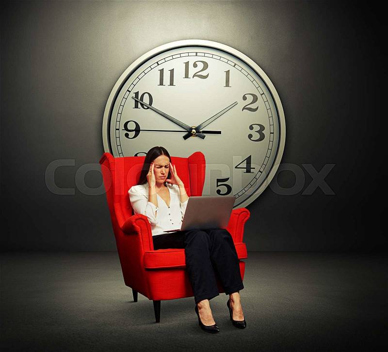 Tired woman with laptop sitting on the red chair over grey wall with big clock, stock photo