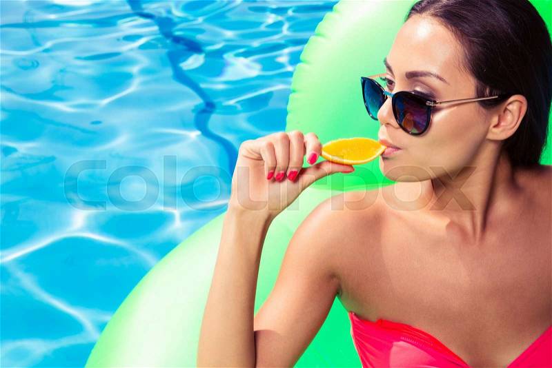 Portrait of a young woman eating orange in swimming pool, stock photo