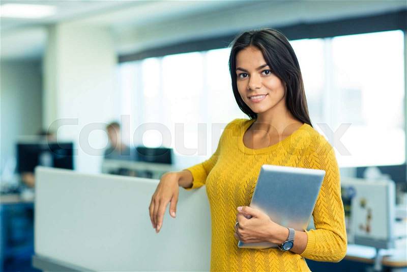Portrait of a smiling cute businesswoman standing with tablet computer in office and looking at camera, stock photo