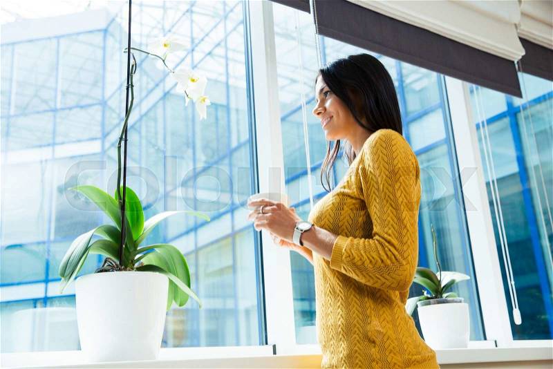 Portrait of a smiling businesswoman holding cup with coffee and looking at window in office, stock photo