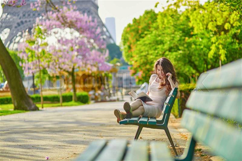Beautiful young woman in Paris, near the Eiffel tower on a nice and sunny spring day, reading on the bench outdoors, stock photo