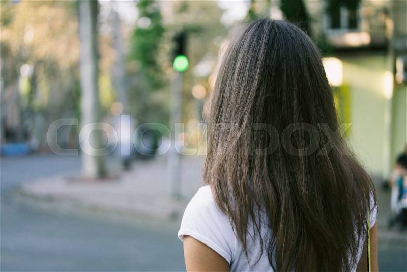 Young female pedestrians cross the road on a green traffic light. The girl has long hair, she is wearing a white T-shirt. Rear view close-up. Woman in the city, stock photo