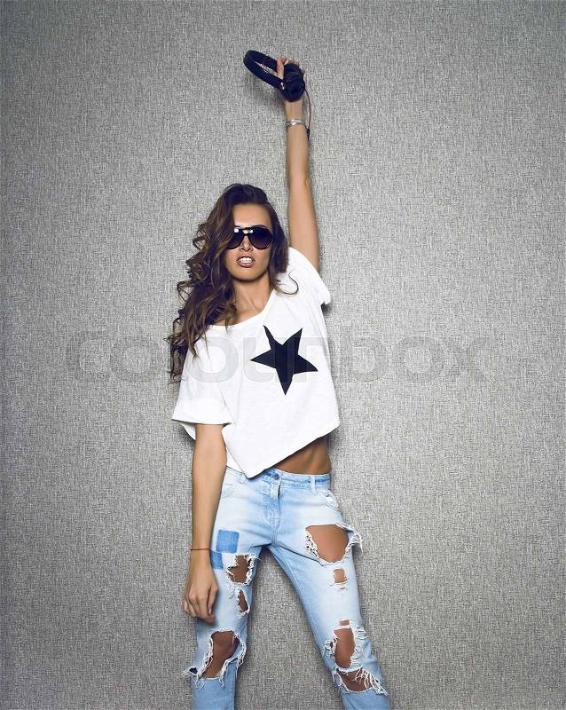 Beautiful dj woman in stylish sunglasses wearing white shirt with black star and blue ripped jeans poses holding black earphones on grey background.Long curly hair.Slim body.Studio shot.Lifestyle.Fashion look.Stylish image.Modern clothes, stock photo