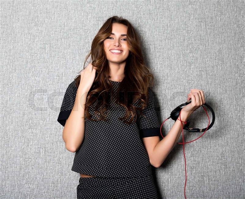 Portrait of beautiful smiling long haired DJ woman with black earphones in dark stylish suit poses on grey background.White teeth.Charming smile.Well-groomed skin.Fashion look.Glamorous clothes.Studio, stock photo