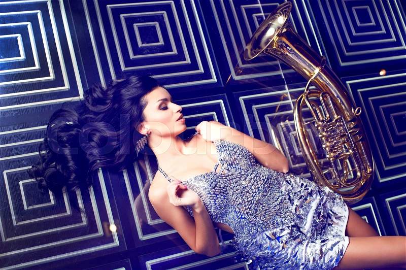 Beautiful elegant woman wearing sparkling shining dress poses in karaoke bar near trumpet.Black long hair.Fashion look.Professional misician.Singer.Stylish image, stock photo