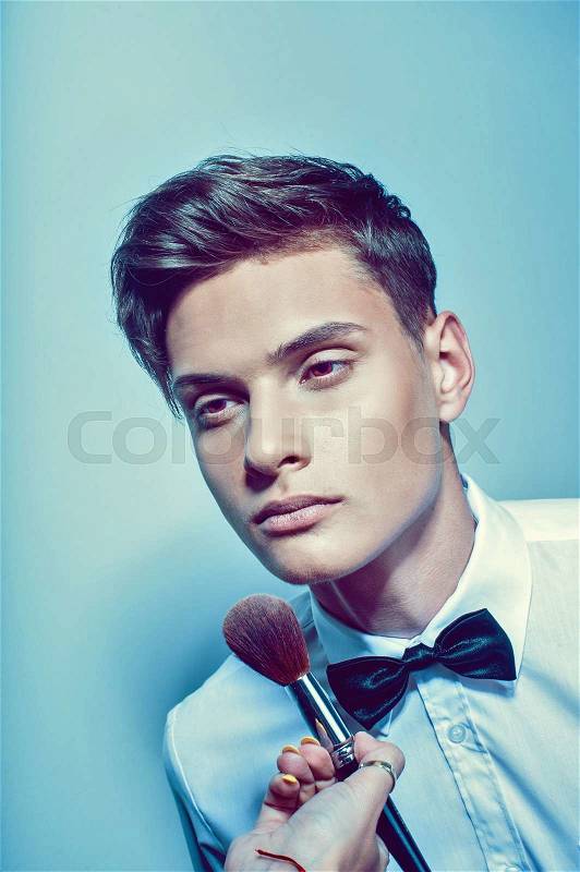 Portrait of beautiful handsome man with tie in white elegant shirt poses on studio background and visagist applyes make up.Fashion look.Cosmetic brush, stock photo