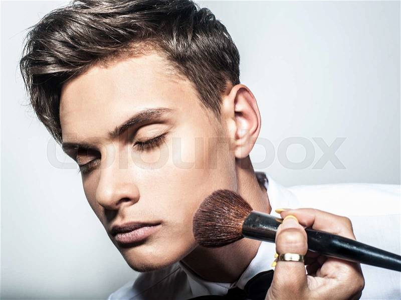 Portrait of beautiful handsome man with tie in white elegant shirt poses on studio background and visagist applyes make up.Fashion look.Cosmetic brush, stock photo