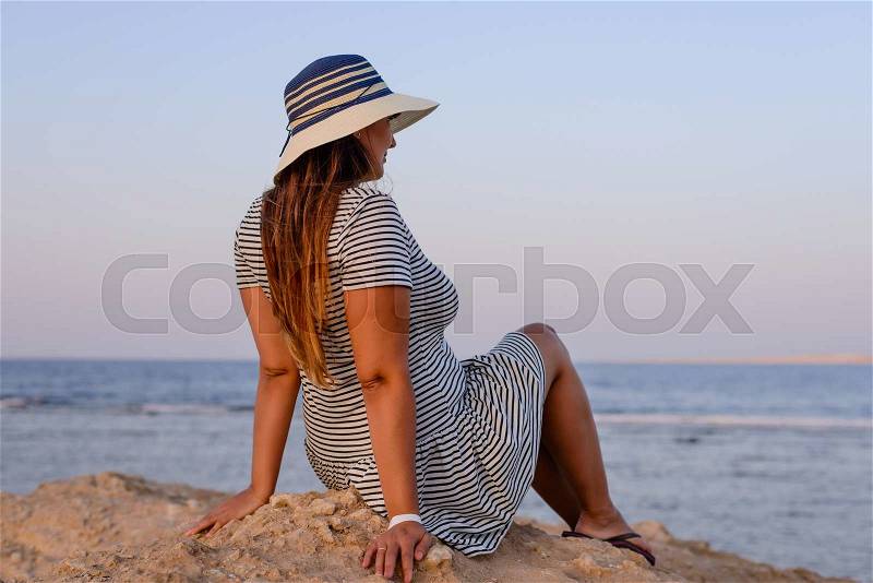 Romantic young woman wearing hat, striped dress and flip flops while sitting on the shore enjoying the breeze at the seaside in the morning, stock photo