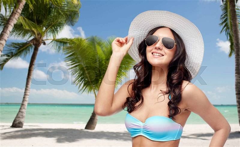People, summer holidays, travel and tourism concept - happy young woman in bikini swimsuit, sunglasses and sun hat over tropical beach with palm trees background, stock photo