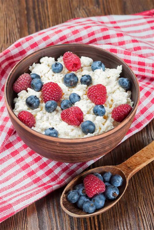Cottage cheese with raspberries and blueberries in brown clay bowl and wooden spoon on red checkered towel , stock photo