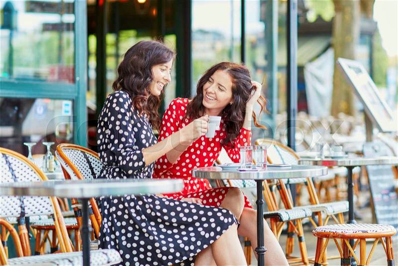 Beautiful twin sisters drinking coffee in a cozy outdoor cafe in Paris, France. Happy smiling girls enjoy their vacation in Europe, stock photo