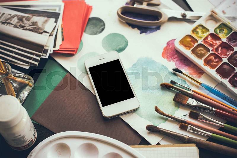 Smartphone on the table in the artist\'s studio, watercolor paints, brushes and sketches, palette and painting tools. Arts and modern technology, hipster style, stock photo