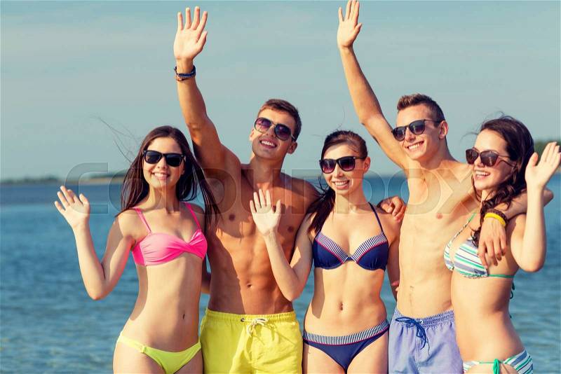 Friendship, sea, holidays, gesture and people concept - group of smiling friends wearing swimwear and sunglasses waving hands on beach, stock photo