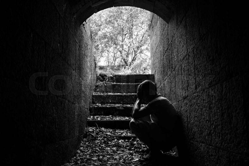 Young sad man sitting inside of dark stone tunnel with stairs in the end, black and white photo, stock photo