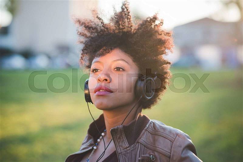 Portrait of a young beautiful black curly hair african woman listening music with headphones in town, overlooking right, pensive - music, relaxing, youth concept, stock photo