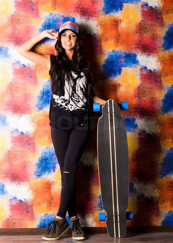 Beautiful smiling brunette woman in color cap,black printed shirt,jeans,leopard shoes poses with longboard sitting on chair. Studio shot.Lifestyle.Healthy life.Sporty girl, stock photo