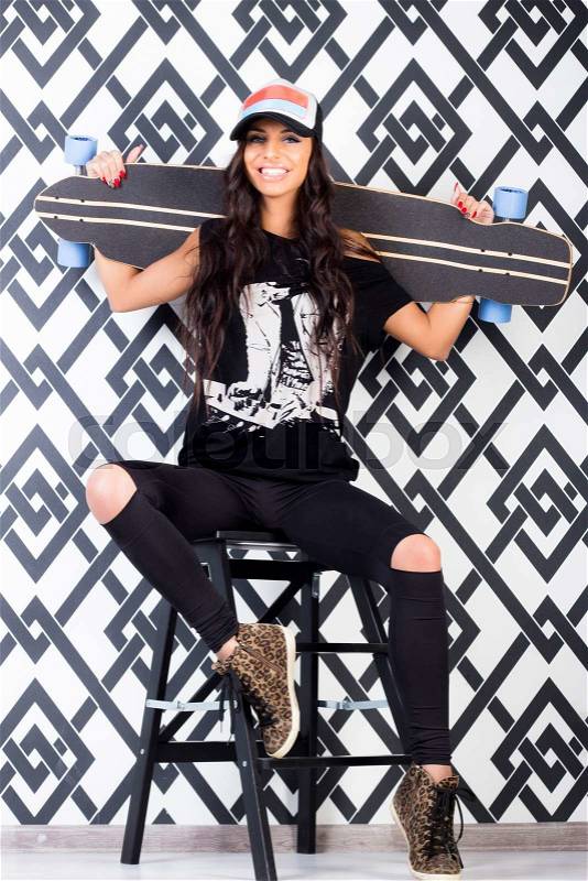 Beautiful smiling brunette woman in color cap,black printed shirt,jeans,leopard shoes poses with longboard sitting on chair. Studio shot.Lifestyle.Healthy life.Sporty girl, stock photo