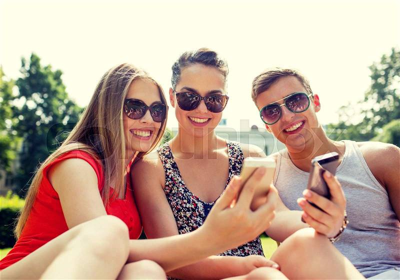 Friendship, leisure, summer, technology and people concept - group of smiling friends with smartphone sitting on grass and making selfie in park, stock photo