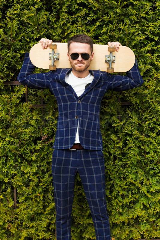 Emotional handsome bearded man in sunglasses and classical suit poses with skateboard on green bush background.Fashion look.Sportrs guy, stock photo